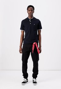 Marineblaues Poloshirt mit weißem Saum, kombiniert mit schwarzen Cargo-Hosen. Rotes Bandana um die Taille gebunden, schwarze Sneaker mit weißen Sohlen.