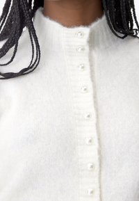 White knit cardigan featuring a ribbed collar and a row of pearl-like buttons down the front. Soft texture and lightweight material.