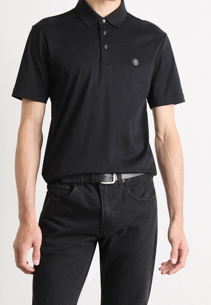 Homme portant un polo noir uni à manches courtes avec trois boutons et un petit logo circulaire sur la poitrine gauche, associé à un jean noir et une ceinture.