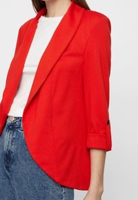 Punainen jakku, jossa on huivikaulus, kolmen neljänneksen hihat ja käännetyt hihansuut. Kangas on sileä ja istuvuus ryhdikäs. Yhdistetty valkoiseen T-paitaan.