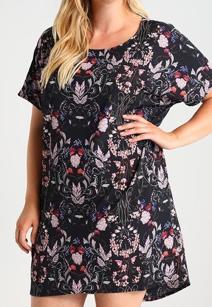 Kobieta ubrana w czarną sukienkę z krótkim rękawem w kwiatowy wzór w kolorach różowym, czerwonym i białym, stoi z jedną ręką na biodrze na jasnym tle.