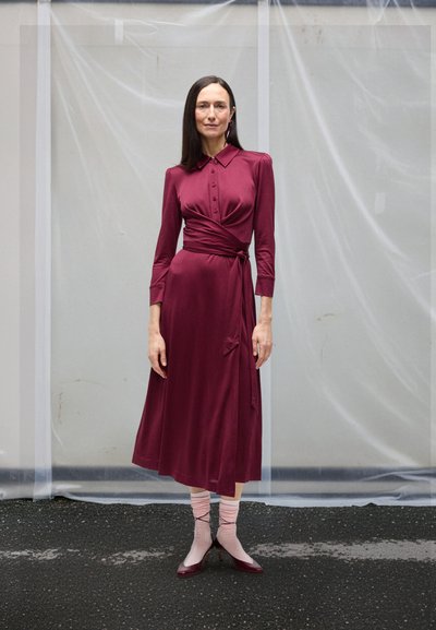 Femme debout dans une robe portefeuille bordeaux à manches longues, portant des chaussettes roses et des talons bordeaux, sur un fond gris uni.