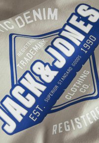 Sivi pamučni majica s plavim i bijelim grafičkim printom s natpisom "JACK & JONES", dizajnom bedža i "EST. 1990" u podebljanim slovima.
