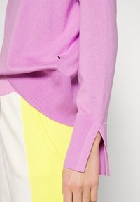 Maglione rosa lavorato a maglia con polsini a coste e slit laterali, abbinato a pantaloni gialli. Il tessuto è liscio e ha una consistenza morbida.