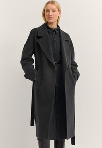 Eine Frau trägt einen dunkelgrauen Wollmantel über einem schwarzen knopfbaren Kleid mit einem Gürtel und steht vor einem schlichten hellen Hintergrund.