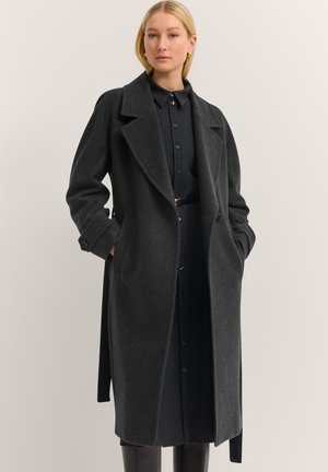 Eine Frau trägt einen dunkelgrauen Wollmantel über einem schwarzen knopfbaren Kleid mit einem Gürtel und steht vor einem schlichten hellen Hintergrund.