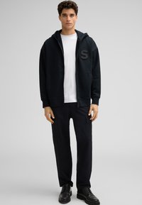 Hoodie zippé noir avec une poche avant et un patch avec la lettre "S" en bold. Porté par-dessus un t-shirt blanc et associé à un pantalon cargo noir et des chaussures noires.
