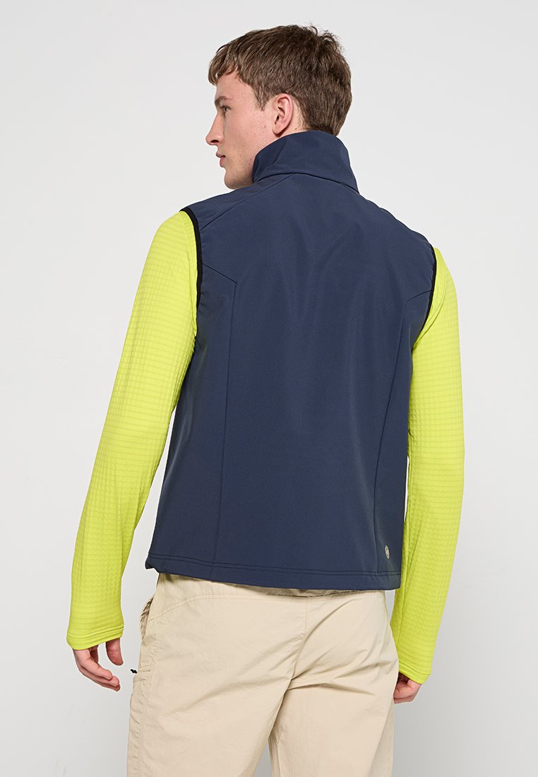 Gilet blu navy realizzato in materiale morbido e resistente all'acqua, con colletto alto. Indossato sopra una camicia a maniche lunghe di un vivace giallo texturizzato. Pantaloni nei toni neutri.