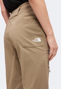 Pantalon beige avec grande poche arrière affichant le logo The North Face porté par une personne avec le bras détendu le long du corps sur un fond uni.