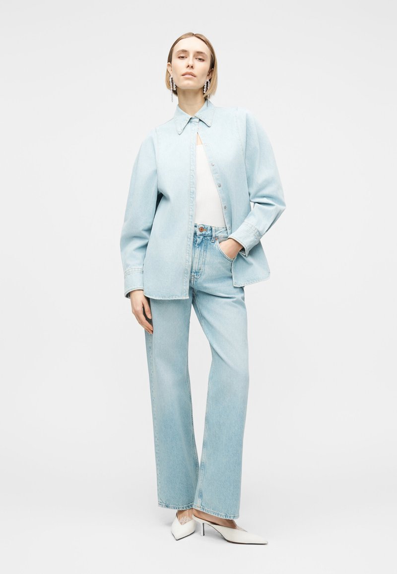 Un mannequin se tient devant un fond blanc, vêtu d’une chemise en jean bleu clair et d’un jean à jambes larges, d’un haut blanc, de boucles d’oreilles pendantes et d’escarpins pointus.