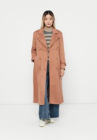 Trench coat lung, cu dublu rând de nasturi, din suede roz deschis, având reveruri largi și buzunare, asortat cu un pulover cu dungi și blugi cu picioare largi.