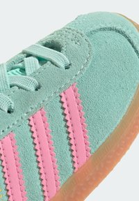 Tenisky ve světle zelené barvě s růžovými cikcak pruhy, strukturovaným semišovým svrškem a průhlednou gumovou podrážkou. Šňůrky ladí s barvou svršku.