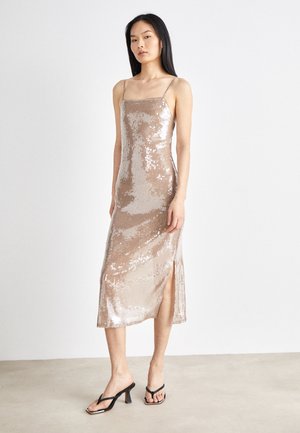 Robe midi en sequins dorés roses avec des fines bretelles et une fente haute sur la cuisse. Texture lisse avec des sequins scintillants sur toute la surface. Portée avec des sandales à talons noires.