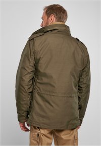 Chaqueta aislante de color verde oliva con un alto cuello, doble bolsillo en el pecho y epaulettes en los hombros. Fabricada con una tela suave y duradera.