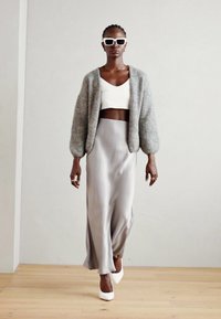 Cardigan gri, pufos, top alb scurt și fustă argintie fluidă. Modelul poartă ochelari de soare albi și pantofi cu toc înalt albi.