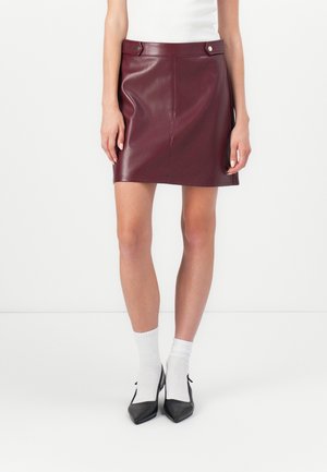 Femme portant une mini-jupe en cuir bordeaux, des chaussettes blanches, des talons noirs pointus et un haut blanc sans manches, debout contre un mur uni.