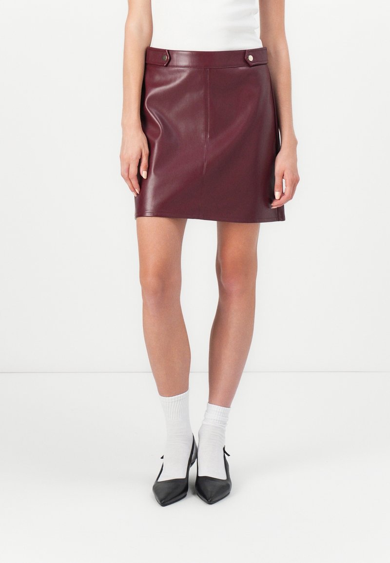 Femme portant une mini-jupe en cuir bordeaux, des chaussettes blanches, des talons noirs pointus et un haut blanc sans manches, debout contre un mur uni.