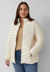 Pufferjacke in Creme mit Reißverschluss, hohem Kragen und diagonaler Steppung. Kombiniert mit einem strukturierten beigen Strickoberteil und blauen Jeans.