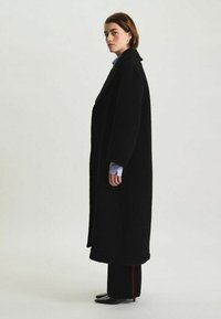 Manteau en laine noire avec des manches larges et une silhouette longue, porté sur une chemise bleu clair à col et un pantalon noir avec une rayure rouge.