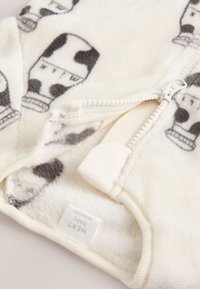 Combinaison pour bébé en polaire couleur crème avec imprimé de bouteille de lait de vache en noir. Fermeture éclair avec bordure en tissu doux près du col.