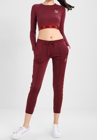 Bordeauxrotes, figurbetontes Crop-Top mit langen Ärmeln und Logo; passende bordeauxrote Jogginghosen, seitliche Akzente in Orange, weiße Sneakers. Glatte Textur.