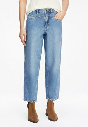 Person wearing light blue straight-leg jeans, standing with one hand in pocket, paired with brown suede ankle boots and white top.