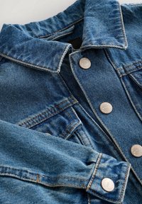 Veste en denim de couleur bleu clair, avec un col classique et des boutons argentés. Présente des accents de couture et une surface en tissu texturé.
