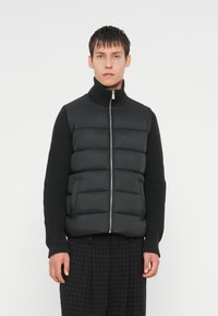 Gilet rembourré noir avec col haut, coutures horizontales et fermeture éclair, associé à un haut en maille noir texturé à manches longues.