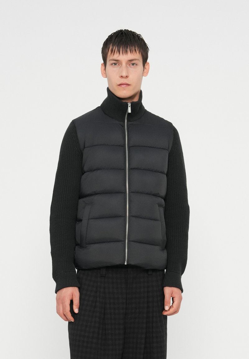 Gilet rembourré noir avec col haut, coutures horizontales et fermeture éclair, associé à un haut en maille noir texturé à manches longues.