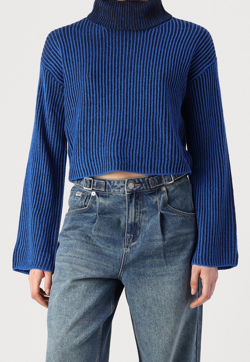 Personne portant un pull court à col roulé rayé bleu et noir avec des manches longues et un jean bleu taille haute.