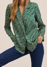 Grüne Satinbluse mit einem Kettenmuster in Weiß und Creme, mit einem eingekerbten Kragen und zwei Vorderaschen. Lange Ärmel.