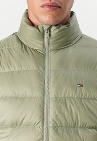 Licht olijfgroene pufferjack met een hoge kraag, een voorrits en horizontaal doorgestikte secties. Kleine logoaccent op de borst.