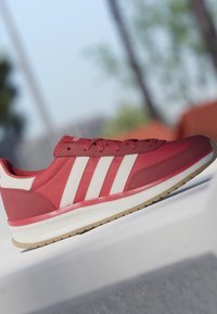 Basket Adidas rouge et blanche avec trois bandes sur le côté, semelle blanche et lacets rouges reposant sur une surface blanche en extérieur.