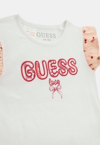 Bijela pamučna majica s crvenim vezom "GUESS love" i mašnom. Ima ružičaste volane na rukavima s uzorkom malih crvenih trešanja.