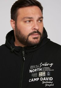 Sudadera negra con cremallera, cordón y texto impreso en blanco, con gráficos de temática marítima y una textura suave y acogedora.