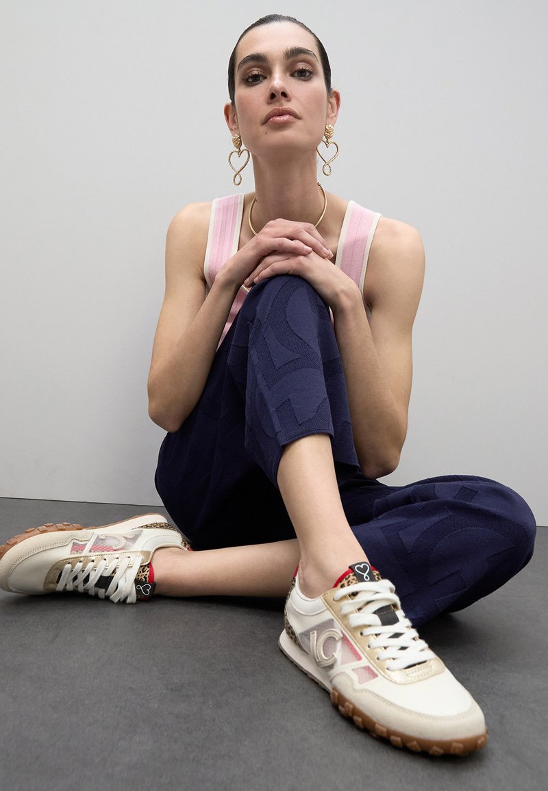 Zapatillas blancas con acentos en beige y rosa, pantalones de tejido azul marino y una blusa rosa clara. Los accesorios incluyen pendientes dorados en forma de corazón.