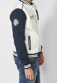 Veste varsity blanche et marine avec des poignets rayés, boutons-pression, poches latérales et logo brodé sur la poitrine et la manche.