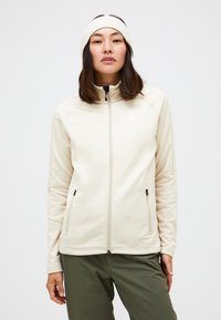 Helle Creme Reißverschlussjacke mit hohem Kragen, glatter Textur, zwei seitlichen Taschen und einem dezenten Logo auf der Brust. Kombiniert mit olivfarbenen Hosen.
