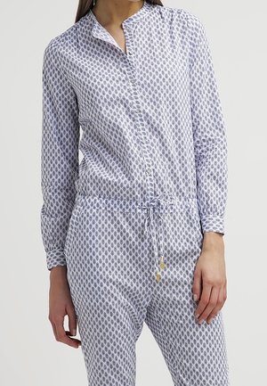 Femme portant une combinaison à manches longues à motif géométrique bleu, resserrée à la taille par un cordon, debout sur un fond uni.