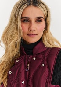 Burgundský prešívaný vest s vysokým golierom, kontrastnými čiernymi texturovanými rukávmi, cvokovými gombíkmi a zipsom s motýlikom ako príveskom.