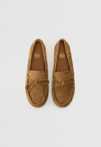 Brown suede moccasin shoes with a rounded toe, lace detailing, and a central bow. Gold-tone hardware accents the sides.