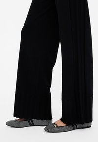 Ballerine con motivo borchiato nero e argento, punta arrotondata e suola piatta, abbinate a pantaloni neri plissettati.