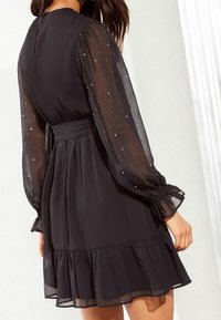 Svart klänning med genomskinliga, långa ärmar prydda med små, silverfärgade prickar. Har en figurnära midja och en volangslå.