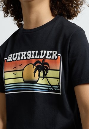 Person iført en sort t-shirt med en farverig solnedgang, palme, fugle og teksten "QUIKSILVER" foran.