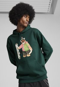 Tamsiai žalia džemperis su grafika, kurioje pavaizduotas lokys rožiniais ir žaliais drabužiais, su akinių nuo saulės ir riedlente. Minkšta medžiaga su kengūros kišene.