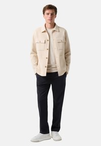 Lys beige uldblandings skjortejakke med to brystlommer og tan knapper, båret over en cremefarvet skjorte, i kombination med marineblå bukser og hvide sneakers.