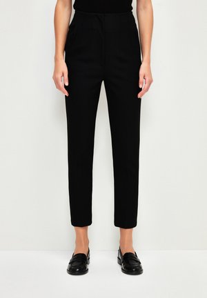 Personne portant un pantalon noir ajusté longueur cheville et des mocassins noirs, debout devant un fond blanc uni.