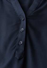 Blusa blu navy con scollo a V, caratterizzata da cinque bottoni rotondi neri e tessuto liscio con una sottile lucentezza. Costruzione e dettagli semplici.