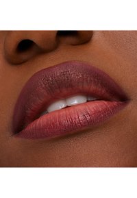 Matte leppestift i dyp plomme påfør på leppene med naturlig glans, som viser en fyldig form og jevn tekstur mot en mørkere hudtone.