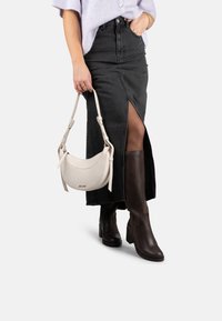 Sac à main en cuir blanc avec une bandoulière ajustable, associé à une jupe en denim noire avec une fente devant, et des bottes en cuir marron jusqu'au genou.